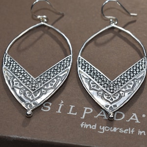 Könnte beinhalten: Ein Paar silberne Ohrringe mit geometrischem Design. Die Ohrringe haben die Form einer Träne mit einer kleinen Perle am unteren Ende. Die Ohrringe befinden sich auf einem braunen Hintergrund mit dem Text "Silpada" und "find yourself in it".