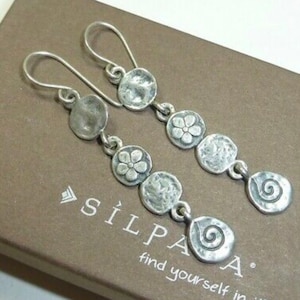 May include: A pair of silver dangle earrings with a floral design. The earrings feature a series of round, textured silver charms, including a flower, a swirl, and a textured disc. The earrings are attached to a silver hook.