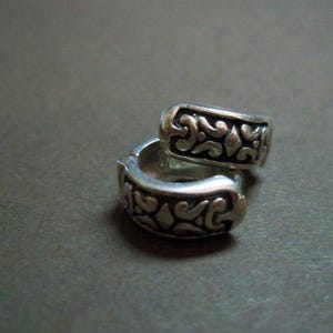 Può includere: Due orecchini a cerchio d'argento con un design ornato e a spirale. Gli orecchini sono impilati l'uno sull'altro, mostrando gli intricati dettagli della lavorazione del metallo. Lo sfondo è grigio.