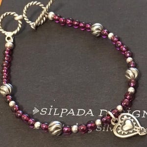 May include: A silver bracelet with a toggle clasp, featuring small, round, purple beads interspersed with silver beads. A silver heart-shaped charm with a textured border hangs from the bracelet. The bracelet is on a dark surface.