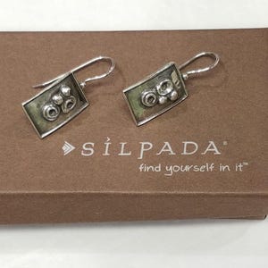 May include: A pair of silver-tone earrings with a rectangular design. Each earring features a hook and a rectangular frame with a textured, green-toned center and circular embellishments. The earrings are displayed on a brown box with the text "SILPADA".
