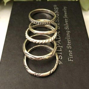 May include: A stack of five silver rings with various designs, including beaded, twisted, and patterned bands. The rings are displayed on a black surface with the text "SILPADA DESIGNS Fine Sterling Silver Jewelry."