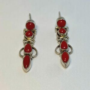 May include: A pair of silver earrings with a dangling design. Each earring features three vibrant red coral stones in a vertical arrangement. The earrings have a post-back closure and are displayed against a white background.