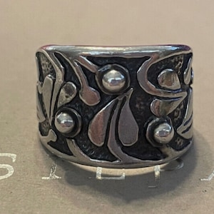 May include: A silver ring with a wide band featuring an intricate design of stylized flowers and leaves. The band is textured and has several raised silver dots.