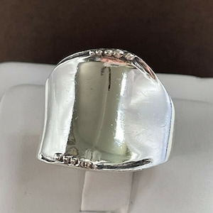 Könnte beinhalten: Ein breiter, polierter Silberring mit glatter, geschwungener Oberfläche. Der Ring hat ein leichtes Wellendesign und ist mit kleinen, funkelnden Verzierungen am oberen Rand versehen. Der Ring wird auf einem weißen Ständer präsentiert.