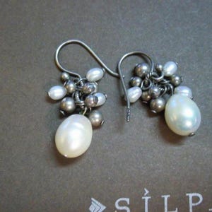 May include: A pair of dangle earrings featuring clusters of small, silver-toned beads and white pearls. Each earring has a large, teardrop-shaped pearl at the bottom. The earrings are on a brown surface.