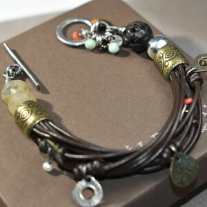 May include: A brown leather bracelet with multiple strands, adorned with gold and silver charms, including a small round charm with the text "self in it".