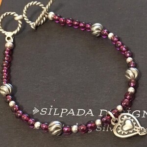 May include: A silver bracelet with a toggle clasp, featuring small, round, purple beads interspersed with silver beads. A silver heart charm with a textured border hangs from the bracelet. The brand name "SILPADA" is visible.
