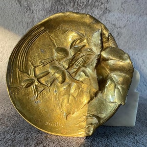 May include: A gold-coloured metal trinket dish with a floral design and the inscription "Detaille" on the bottom.