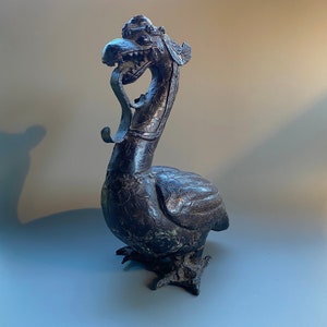 Puede incluir: Una escultura de bronce negro de un pájaro mítico con cuello largo y cabeza detallada. El pájaro tiene un pico largo y curvo y una corona en la cabeza. La escultura está de pie sobre una superficie plana.