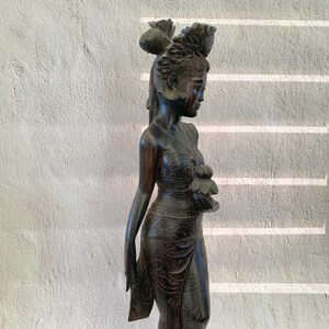 Puede incluir: Una estatua de madera oscura tallada de una mujer con un tocado tradicional. La estatua está de pie con una mano en la cadera y la otra sosteniendo una flor.