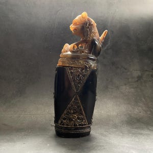 Puede incluir: Un recipiente de cuerno tallado con un diseño detallado. El recipiente es negro con tallas intrincadas y una figura de león dorada en la parte superior.