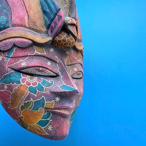 Könnte beinhalten: Eine Holzmaske mit aufwendigen floralen Mustern in Rot-, Blau-, Grün- und Gelbtönen. Die Maske hat einen Ausdruck mit geschlossenen Augen und eine geschnitzte Nase.