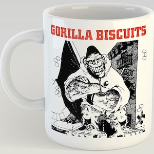 May include: White ceramic mug with a black and white gorilla graphic wearing a hoodie, and the red text "GORILLA BISCUITS". The mug has a curved handle and a cylindrical shape, suitable for hot beverages.