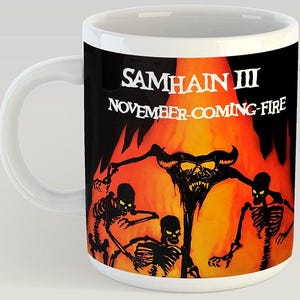 May include: White ceramic mug featuring a black and orange graphic with the text "SAMHAIN III NOVEMBER-COMING-FIRE." The design includes skeletal figures and a horned demon against a fiery background.