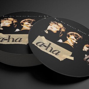 Op de afbeelding: Zwarte en witte ronde onderzetters met het logo van de band a-ha en de tekst "The Sun Always Shines On T.V." erop gedrukt.
