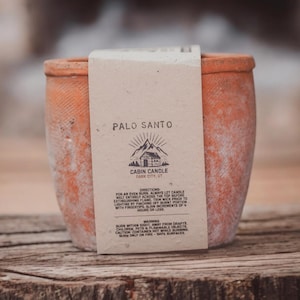 May include: A terracotta candle jar with a label that reads "Palo Santo Cabin Candle Park City, UT". The label also includes directions for burning the candle and warnings.