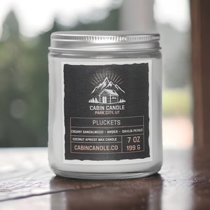 May include: A white candle jar with a black label. The label features a cabin illustration and the text "CABIN CANDLE PARK CITY, UT" and "PLUCKETS". The candle is a coconut apricot wax candle with the scent of creamy sandalwood, amber, and dahlia petals. The jar is 7 ounces or 199 grams.