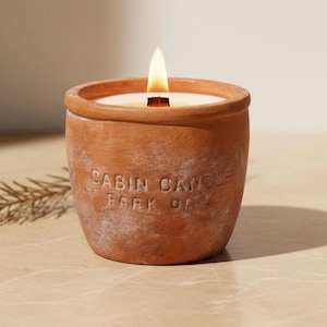 May include: A lit candle in a terracotta pot. The candle has a wooden wick and is inscribed with the words "CABIN CANDLE PARK OK". The pot is a warm, earthy orange color.
