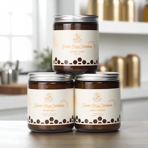 May include: Three glass jar candles with silver lids are stacked. Each jar has a brown label with the text "Desert Hive Collection" and a honeycomb pattern. The labels also read "Natural Honey", "Nature Walk", and "Lemon Lavender".