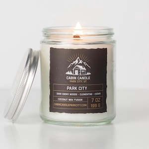 May include: A lit glass jar candle with a silver lid. The candle has a black label with white text that reads "CABIN CANDLE PARK CITY, UT" and "PARK CITY". The candle is 7 oz and 199 g.