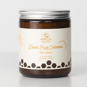 May include: A brown glass jar candle with a silver lid. The label reads "Desert Hive Collection" and "Palo Santo." The label also includes the text "Palo Santo | Sage | Cedarwood | Orange" and a mountain and cabin logo. The candle is made with Utah's pure beeswax.