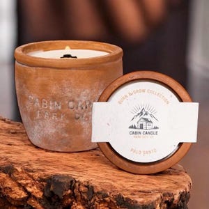May include: A lit, white candle in a terracotta pot with the text "CABIN CANDLE PARK CITY UT". The candle sits on a wooden surface next to a wooden lid with the text "BURN & GROW COLLECTION" and a mountain cabin logo.