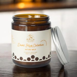 May include: A brown glass jar candle with a silver lid, labeled "Desert Hive Collection Nature Walk." The label includes text about the scent: Fir Needles, Earthy Moss, Amber, and Eucalyptus. The jar is on a wooden surface.