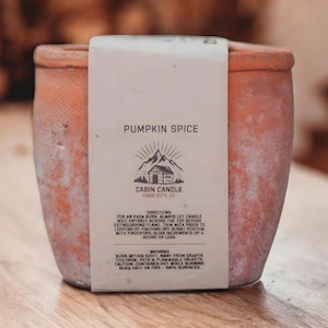 May include: A terracotta candle jar with a white label that reads "Pumpkin Spice Cabin Candle Park City, UT". The label also includes directions for burning the candle and a warning.