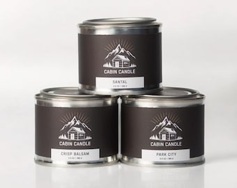 May include: Three metal tin candles stacked on top of each other. Each candle has a dark brown label with a white mountain and cabin logo, and the words "Cabin Candle". The bottom candles read "Crisp Balsam" and "Park City". The top candle reads "Santal".