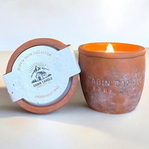 May include: A lit grapefruit pine scented candle in a terracotta pot. The pot has the words "Cabin Candle Park City" embossed on the side. The lid is off, revealing the candle and a label with the text "Burn & Grow Collection".
