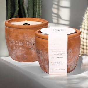 May include: Two terracotta-colored candles in a rustic style. One candle is lit, and the other has a tag that reads "PLANT THIS TAG". The candles are in a pot shape, with the text "CABIN OAK FARM CO." on one.