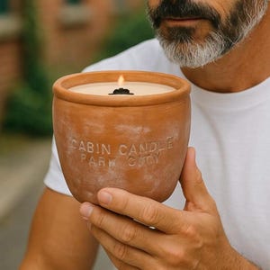 May include: A lit, terracotta-colored candle in a round pot. The candle has a light-colored wax and a black wick. The pot has the words "CABIN CANDLE PARK CITY" embossed on the side. The candle is held by a person wearing a white t-shirt.
