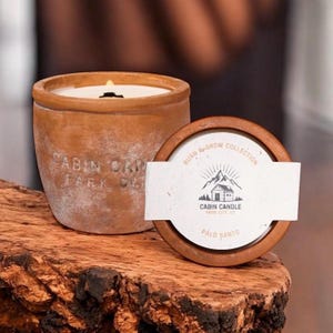 May include: A lit Palo Santo scented candle in a terracotta pot. The pot is labeled "CABIN CANDLE PARK CITY UT". The candle sits next to its wooden lid, which has the text "BURN & GROW COLLECTION" and a logo.