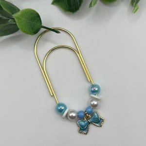 May include: A large gold-toned metal paperclip embellished with a decorative charm arrangement. The beads include iridescent light blue spheres, solid white spheres, and small light blue spacers, finished with a light blue enamel bow charm.