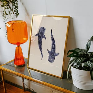 K&ouml;nnte beinhalten: Gerahmter Kunstdruck mit zwei blau-wei&szlig;en Walhaien auf cremefarbenem Hintergrund. Der Druck befindet sich in einem goldenen Rahmen und steht auf einem Tisch mit einer orangefarbenen Lampe und einer Topfpflanze.