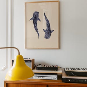 K&ouml;nnte beinhalten: Ein gerahmter Kunstdruck mit zwei stilisierten Walhaien in Marineblau vor beigem Hintergrund. Das Kunstwerk ist in einem hellen Holzrahmen gefasst, ideal f&uuml;r die Inneneinrichtung. Der Druck wird an einer Wand &uuml;ber einem Holzschrank pr&auml;sentiert.