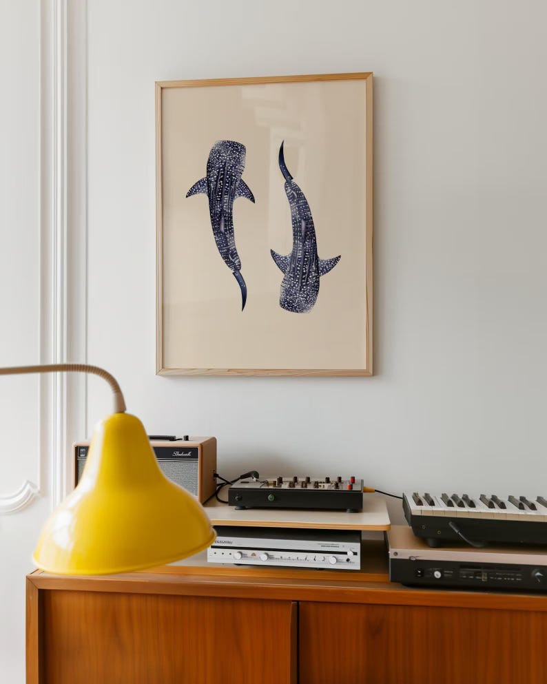 K&ouml;nnte beinhalten: Ein gerahmter Kunstdruck mit zwei stilisierten Walhaien in Marineblau vor beigem Hintergrund. Das Kunstwerk ist in einem hellen Holzrahmen gefasst, ideal f&uuml;r die Inneneinrichtung. Der Druck wird an einer Wand &uuml;ber einem Holzschrank pr&auml;sentiert.
