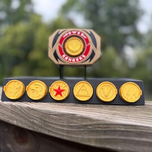 Puede incluir: Una exhibición de Power Rangers con un morpher dorado y negro con las palabras "POWER RANGERS" y seis monedas de oro con diferentes símbolos. La exhibición está sobre una base negra, sobre un fondo verde borroso.