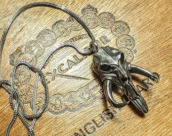 May include: A silver chain necklace with a detailed skull pendant. The pendant features a skull with horns and a textured surface. The necklace is displayed on a wooden box with the words "English Cigar" printed on it.
