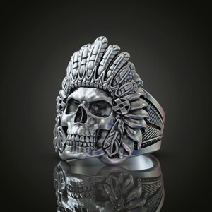 May include: A silver ring featuring a skull wearing a headdress of feathers. The ring is intricately detailed and has a textured finish.