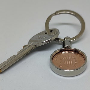May include: A silver key chain with a key and a copper-colored coin featuring a building with a crown on top. The coin is surrounded by a silver ring.