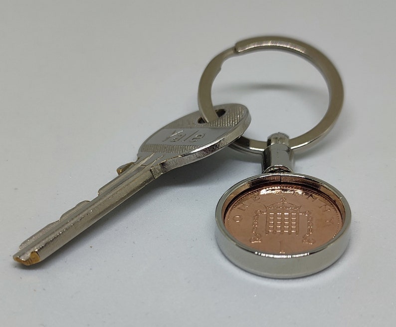 May include: A silver key chain with a key and a copper-colored coin featuring a building with a crown on top. The coin is surrounded by a silver ring.