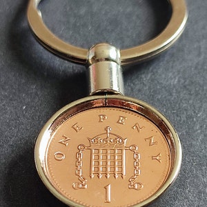 May include: A silver key chain with a one penny coin attached. The coin features a depiction of the Houses of Parliament and the inscription "ONE PENNY".