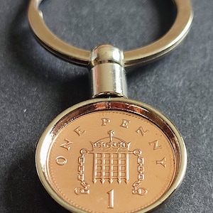 May include: A silver key chain with a one penny coin attached. The coin features a depiction of the Houses of Parliament and the inscription "ONE PENNY".