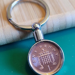 May include: A silver key chain with a one penny coin attached. The coin is copper colored and features the British coat of arms with the words "ONE PENNY" around the edge.