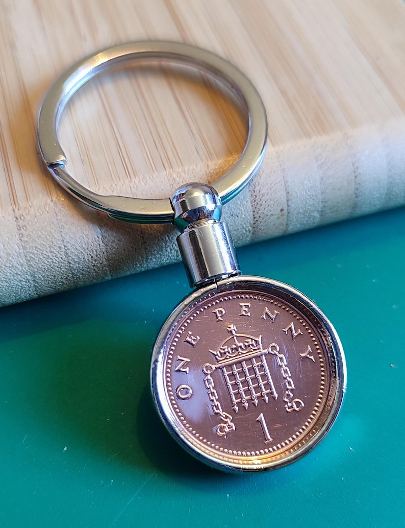 May include: A silver key chain with a one penny coin attached. The coin is copper colored and features the British coat of arms with the words "ONE PENNY" around the edge.