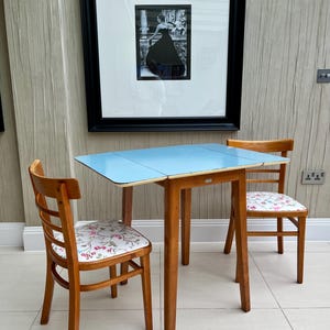 May include: A small, vintage dining set with a light blue tabletop and two wooden chairs. The chairs have floral patterned seat cushions. A framed black and white photograph hangs on the wall behind the table.