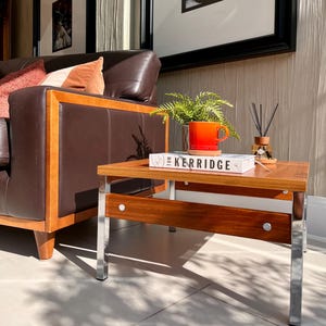 May include: A rectangular coffee table with a wooden top and chrome legs. An orange mug, a book titled "Kerridge", and a small plant are on the table. A brown leather sofa is in the background. The table is approximately 90 cm long.