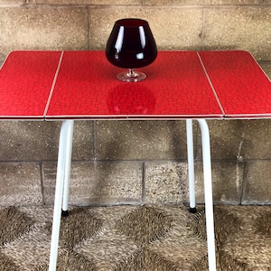 May include: A vintage red table with a patterned surface and white legs. A large, dark red glass sits on the table. The table is approximately 76 cm long and 51 cm wide.